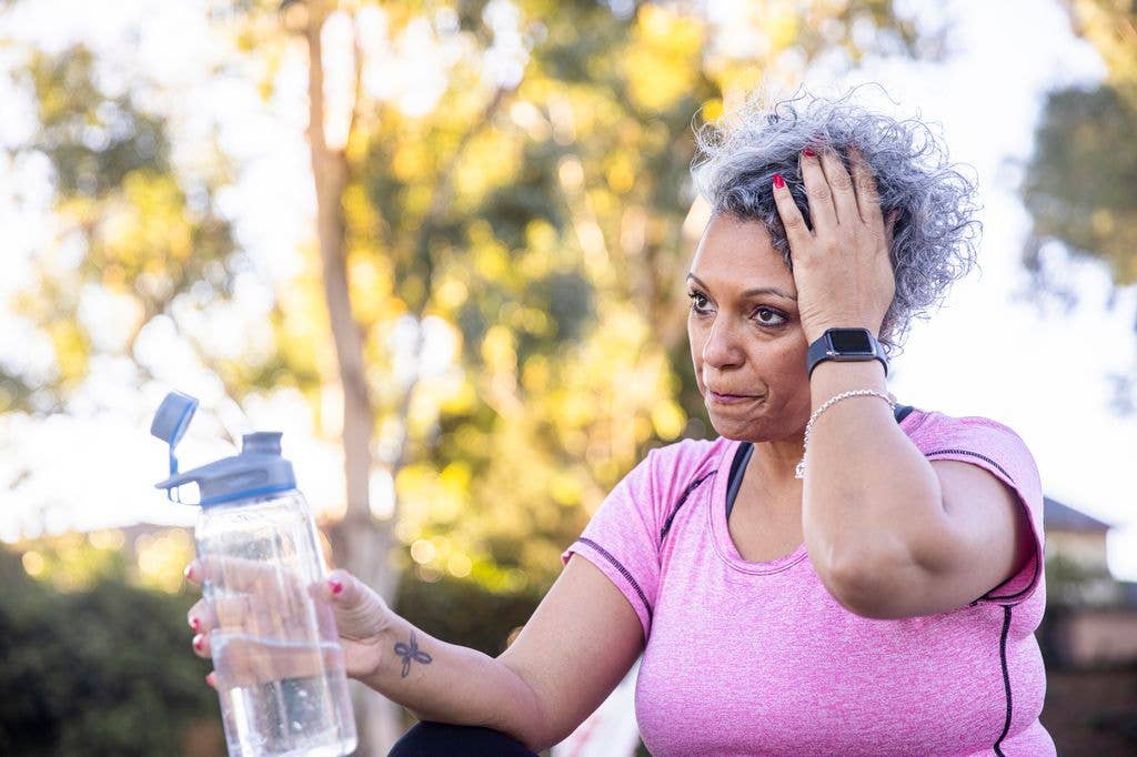 A woman with gray hair holds a water bottle, touching her head with a concerned look.