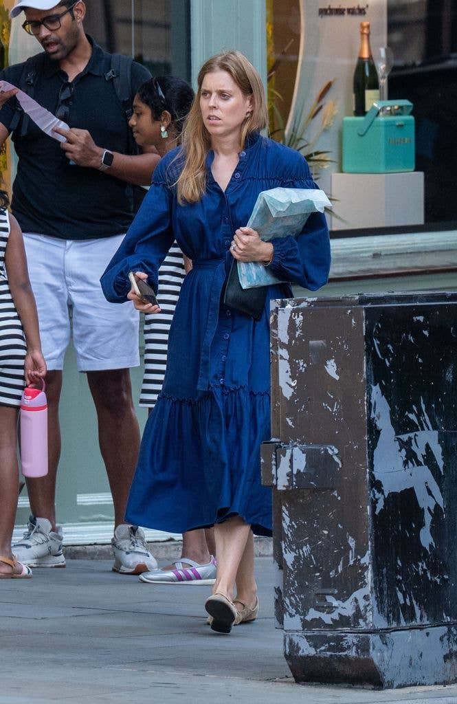 A woman in a blue dress walks down the street with a man and a girl. She holds a plastic bag and a cell phone.