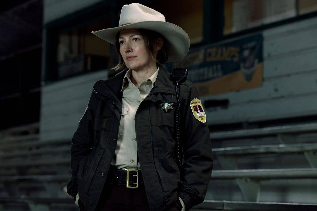 Kelly Macdonald stands in a stadium wearing a black jacket and white hat.
