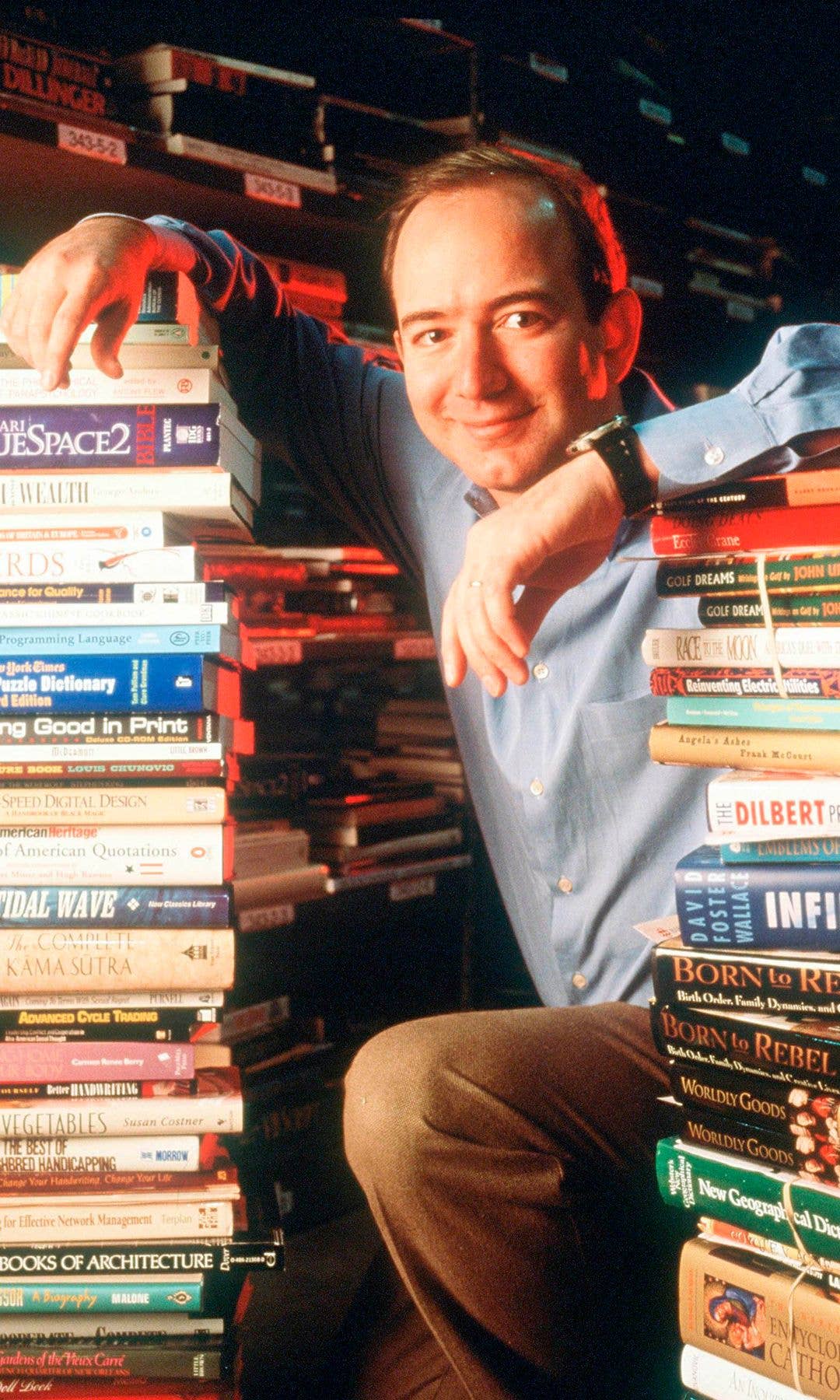 Jeff Bezos, smiling, sits with a stack of books, wearing a blue shirt and brown pants.
