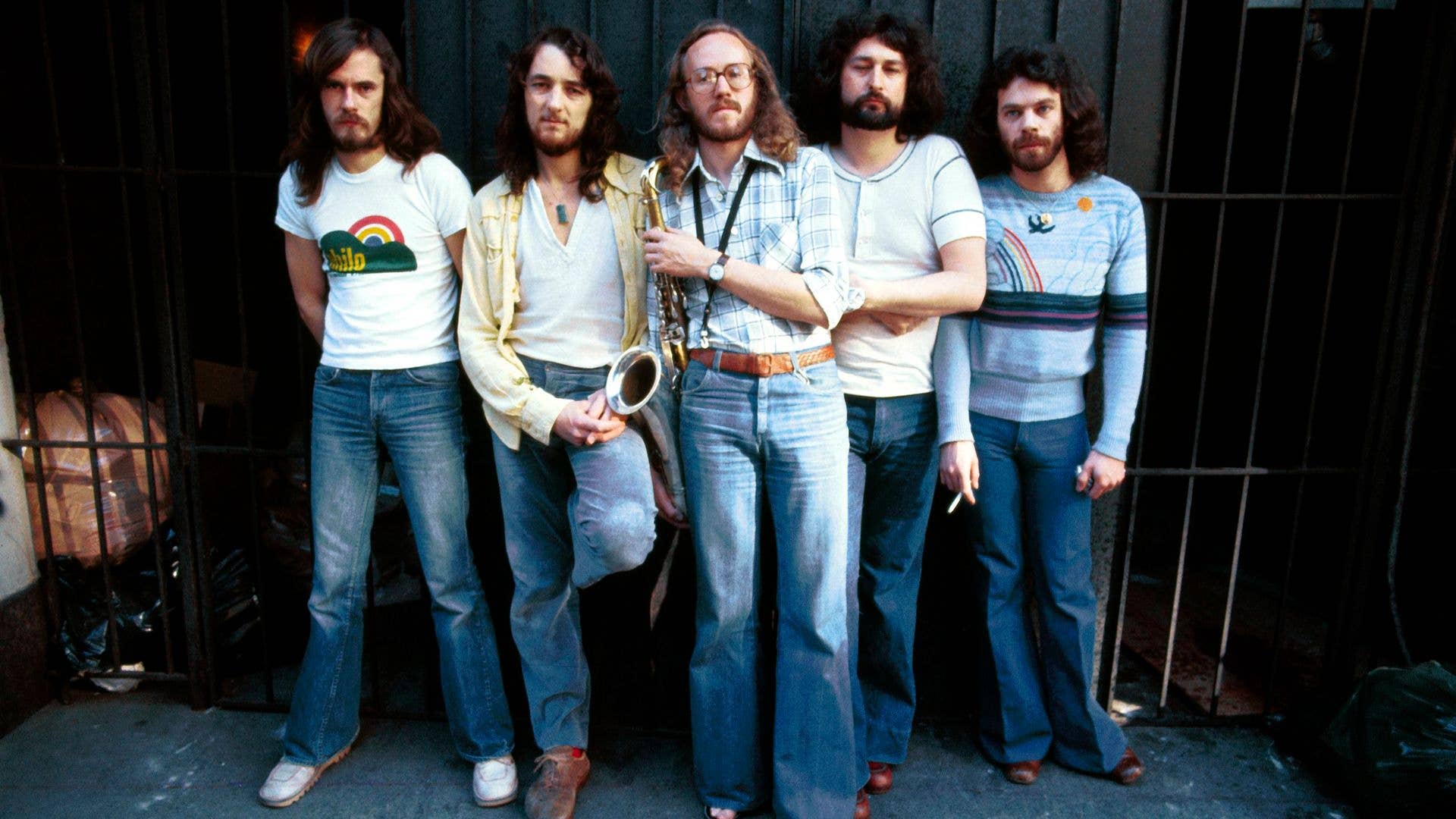 Five men stand together, one holds saxophone; varied clothing, casual setting.