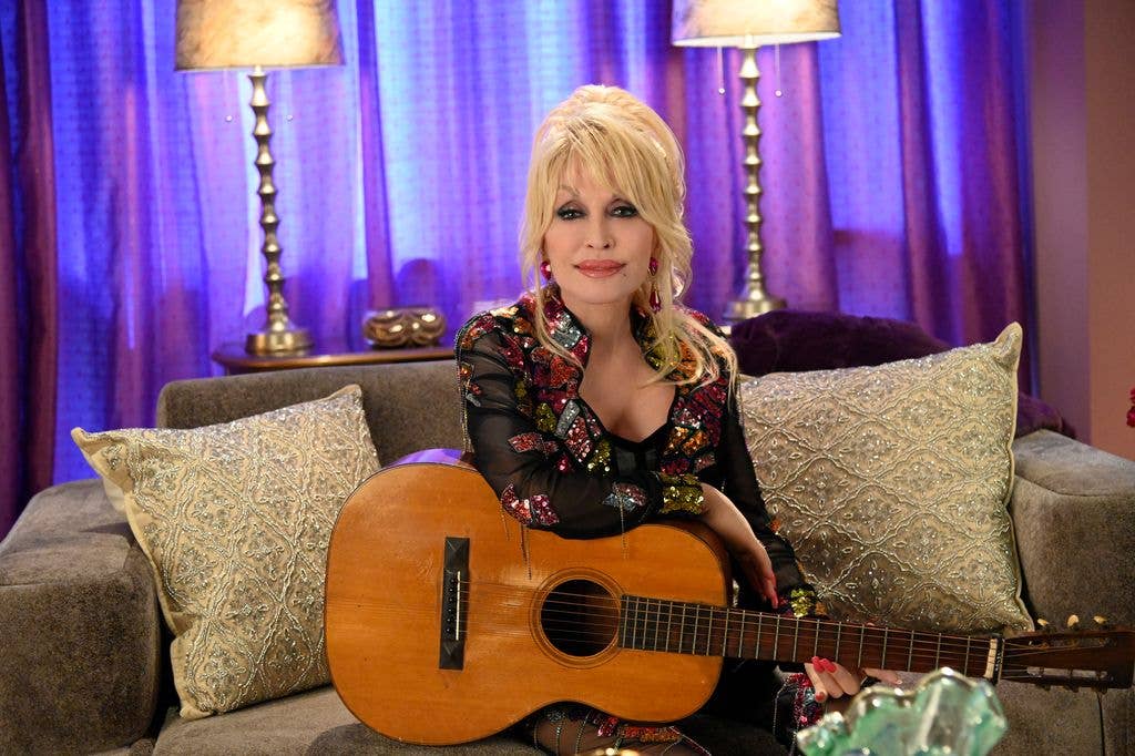 Dolly Parton sitting on a grey sofa with her arm resting on an acoustic guitar.