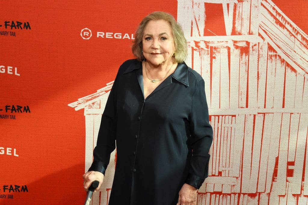 Kathleen Turner stands with a cane, wearing a black blouse, in front of a red and white backdrop.