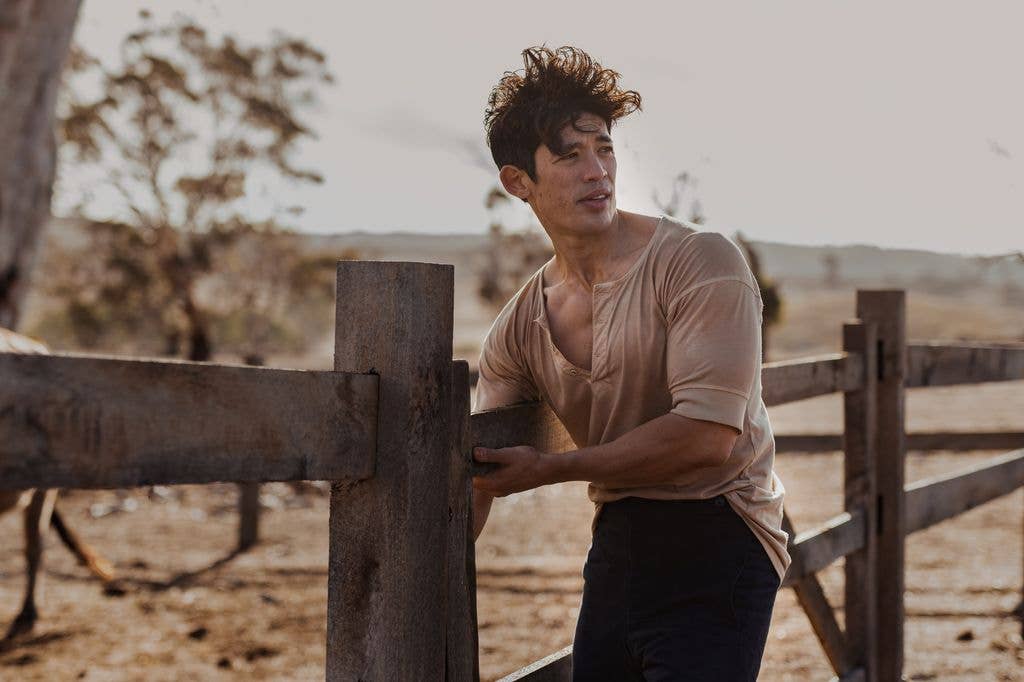 A man (Christopher Chung) with short hair stands by a wooden fence in a rural setting, wearing a beige shirt and dark pants.