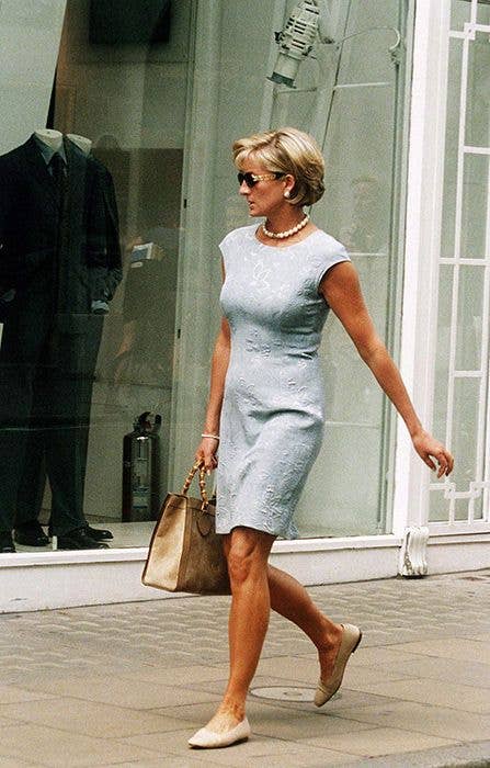 Diana, Princess of Wales, wearing a light blue dress and sunglasses, walks down a sidewalk carrying a handbag.