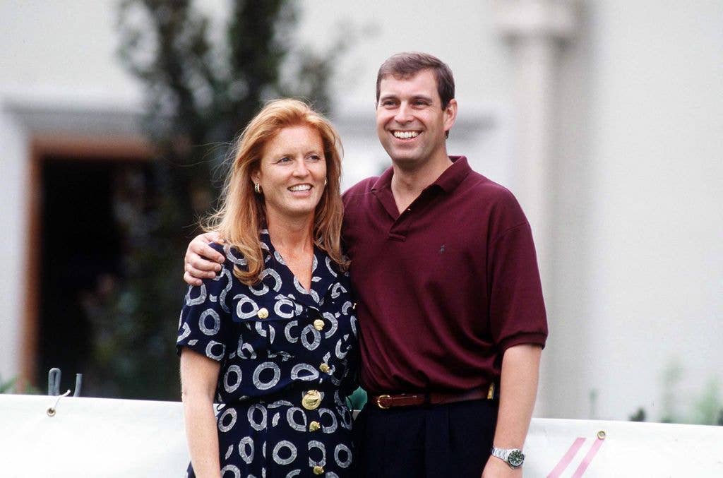 Sarah Ferguson smiling wearing a patterned navy dress and Andrew Mountbaten-Windsor with his arms around her smiling wearing a red shirt