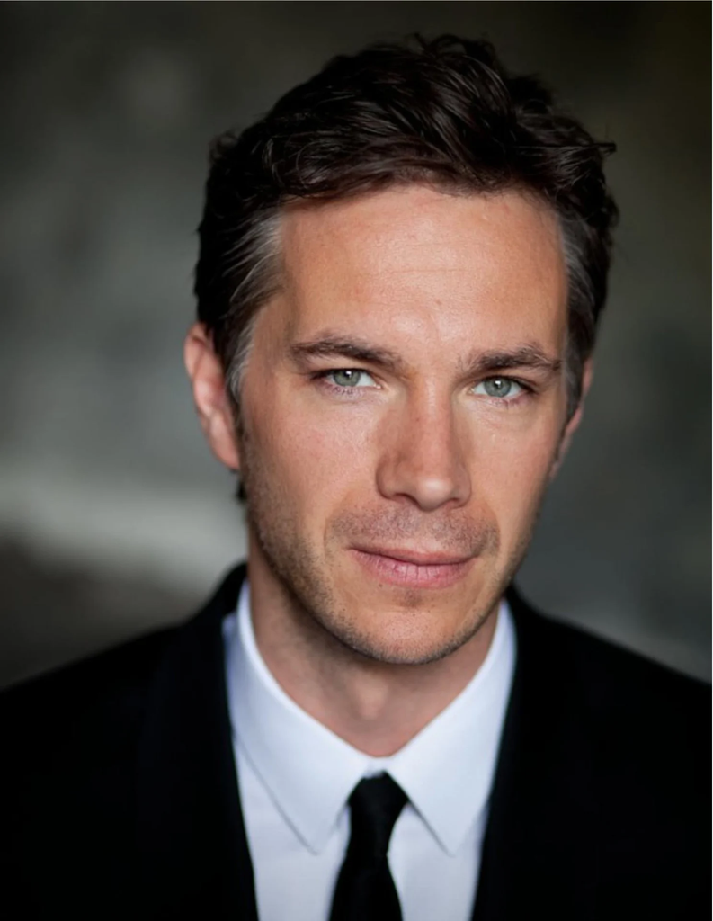James D'Arcy in a black suit and white shirt with a black tie, smiling.
