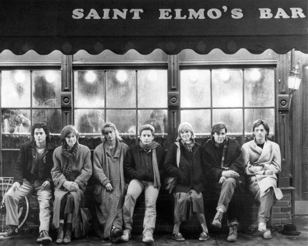 Seven individuals, including Judd Nelson, Emilio Estevez, and Rob Lowe, sit outside Saint Elmo's Bar, dressed in winter clothing.