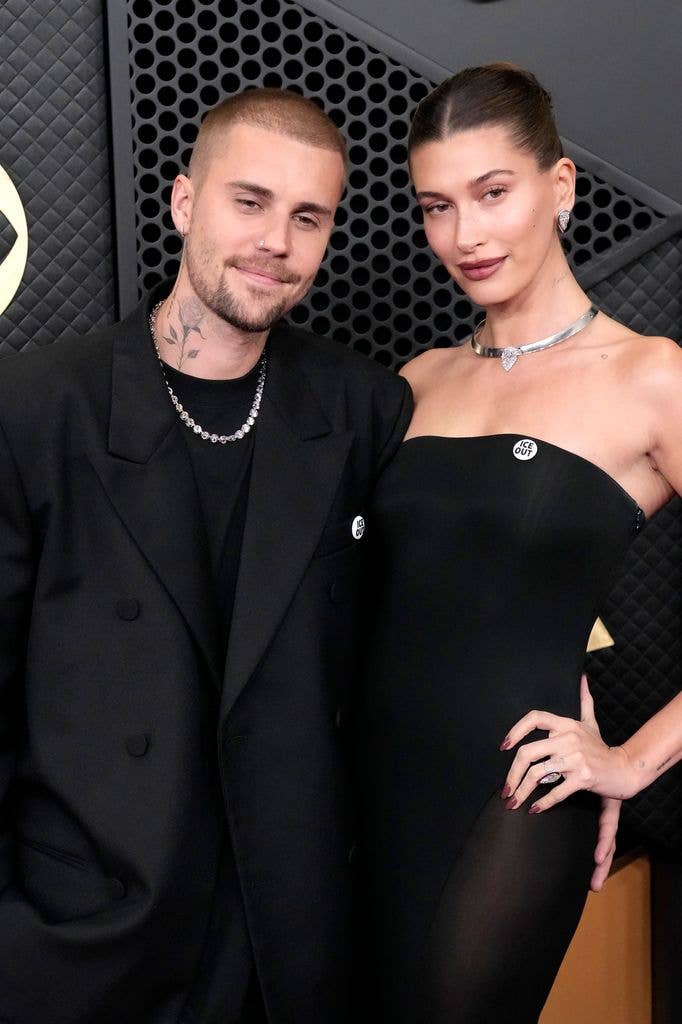 Justin Bieber and Hailey Bieber hold an arm around one another. Justin wears a black suit with a silver necklace and a small 'ICE OUT' pin. Hailey holds her left hand on her hip, wearing a strapless black dress with a silver choker necklace and a white pin with 'ICE OUT' printed in black.