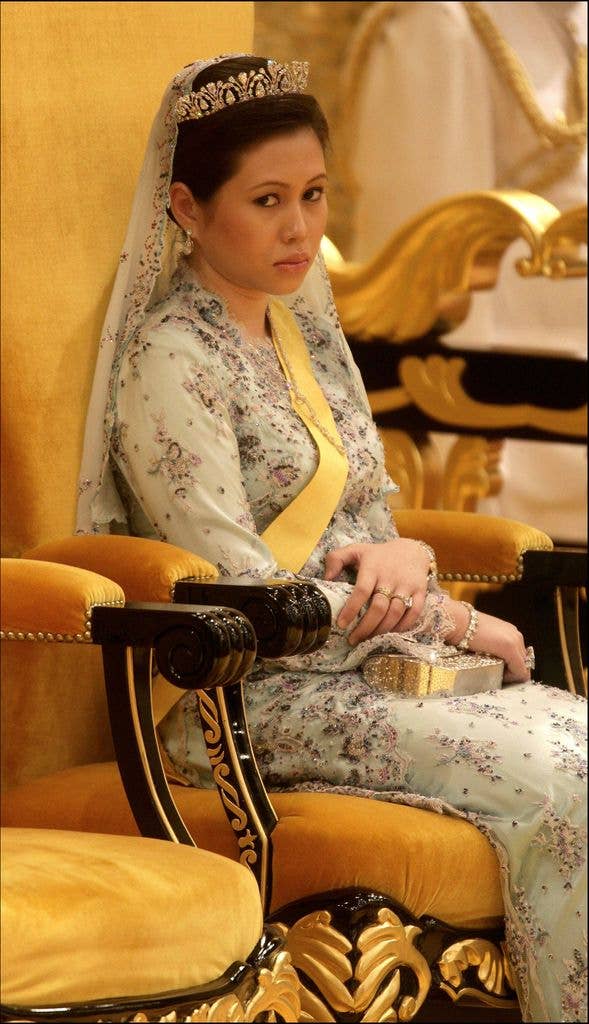 A woman in a floral gown and tiara sits on a golden throne, holding a clutch.