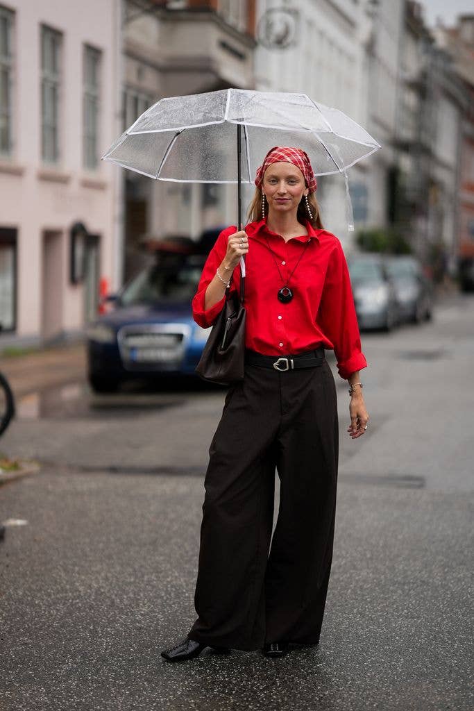Laura Schulte is seen wearing a red long sleeve button up shirt with rolled sleeves; a black cord necklace with a round pendant; dark brown wide leg trousers with a black leather belt; black square toe leather shoes; a dark brown leather shoulder bag; silver drop earrings, silver bracelets and silver rings; a transparent umbrella; blonde hair worn loose beneath a red plaid headscarf during Day Two of the Copenhagen Fashion Week Spring/Summer 2027, outside the MKDT show, on August 4, 2026 in Copenhagen, Denmark. (Photo by Moritz Scholz/Getty Images)