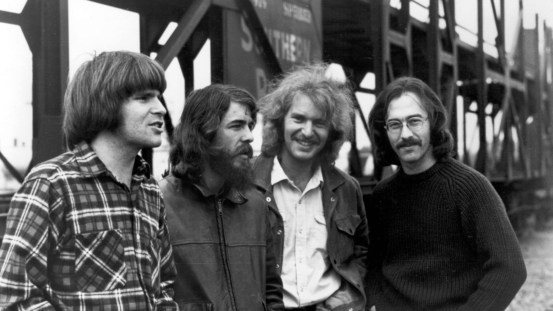 Four men stand together near a bridge. From left: man in plaid shirt, bearded man, smiling man in white shirt, and man with glasses and mustache.