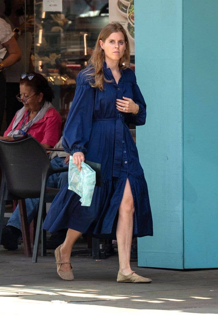 Princess Beatrice in a blue dress, holding a blue pouch, walking outside a store.