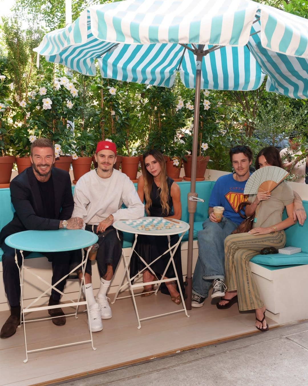 David Beckham, man in red hat, woman in black dress, man in blue shirt, and woman with fan sit under umbrellas.