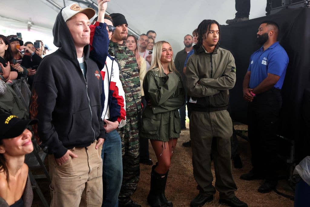 Group of people in a crowd, one man in a camo jacket, another in a green jacket, and a woman with blonde hair.
