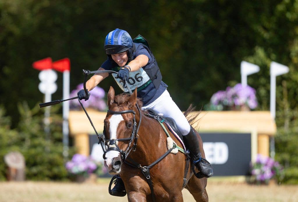Zara Tindall riding a brown horse, holding a stick, wearing a helmet and gloves.