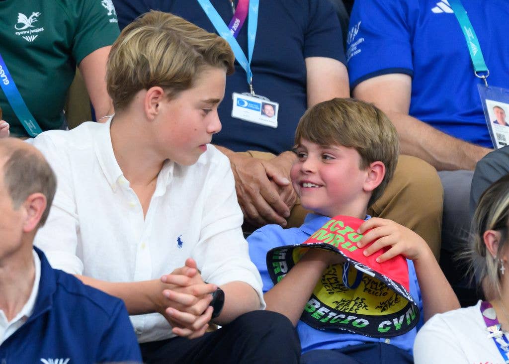 Two young boys, one in a white shirt and the other in a blue shirt with a red cap, sit among a crowd of spectators.