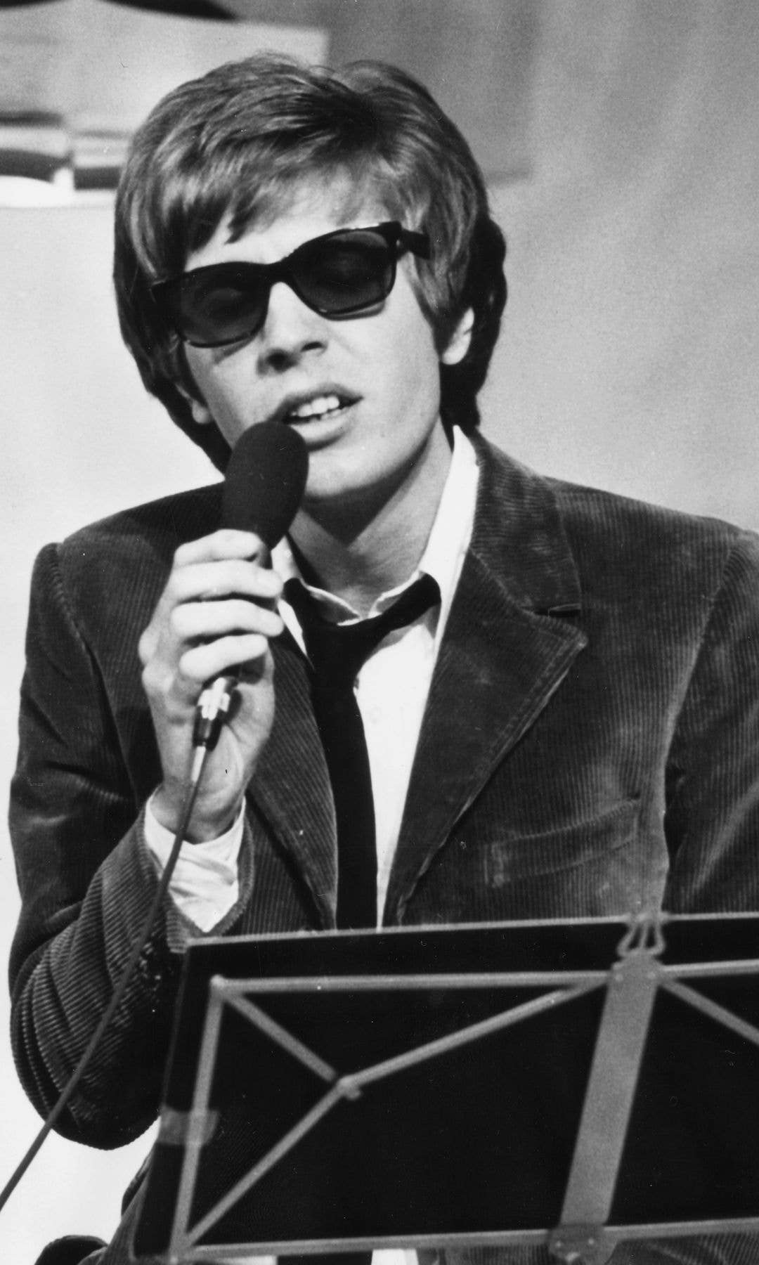 A man in a suit and sunglasses holds a microphone, standing by a music stand.