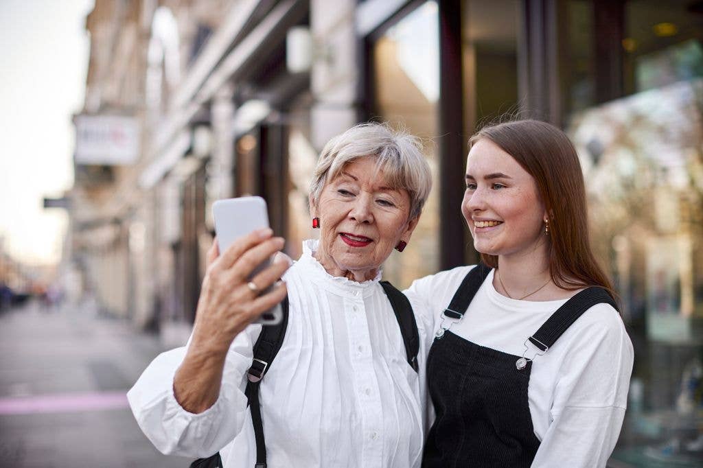 Two females stand on a city street, one taking a selfie with a smartphone. The older woman wears a white blouse, and the teen wears black overalls over a white shirt.