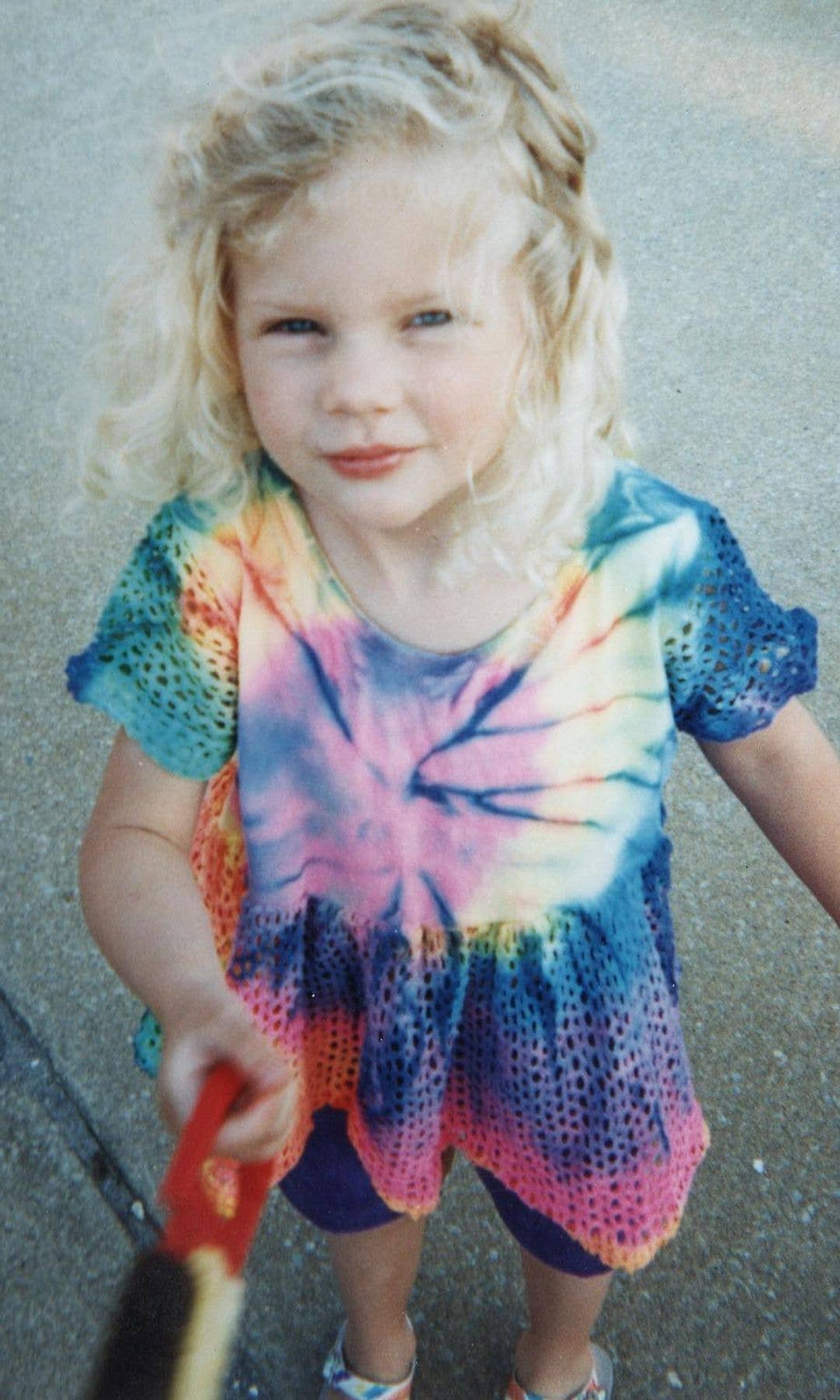 Young girl with blonde hair in a tie-dye dress holding a red object.