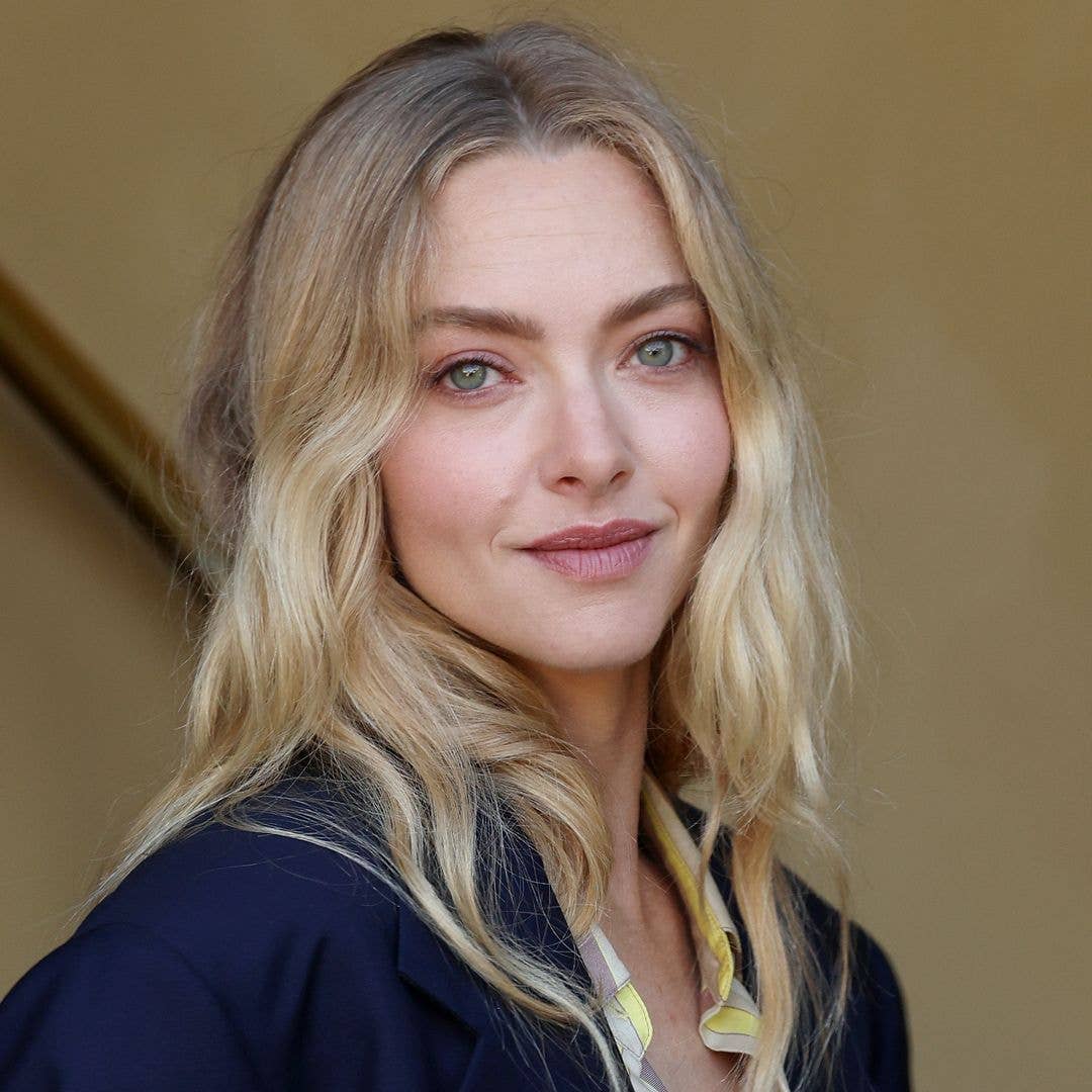 Amanda Seyfried stands in a hallway, wearing a blue blazer and striped shirt, smiling.