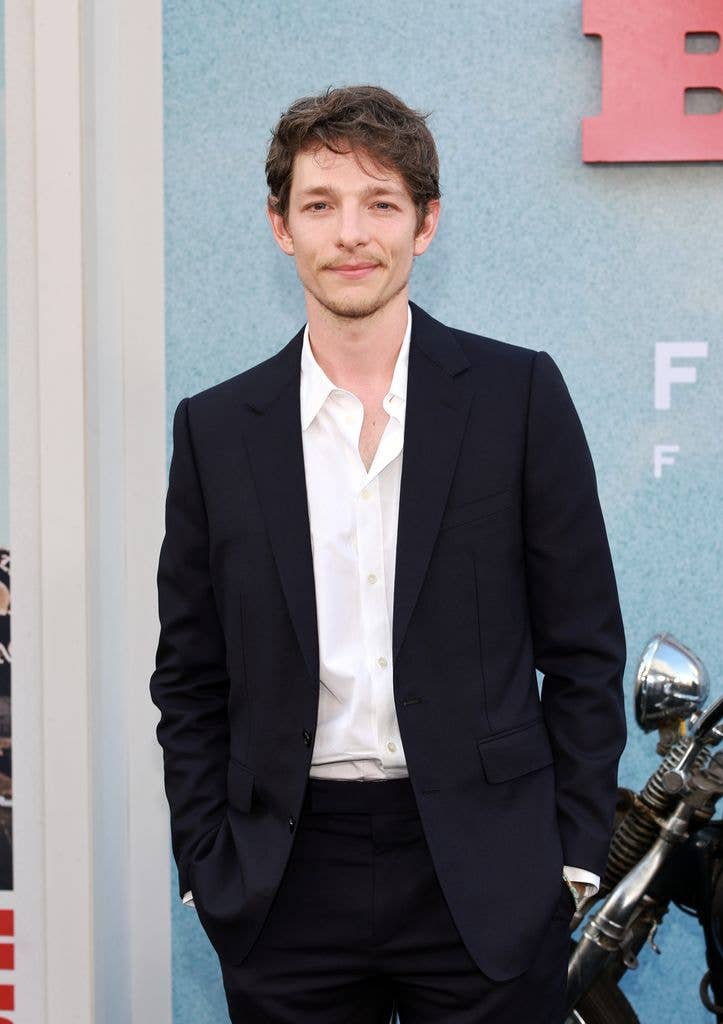 Mike Faist in black suit and white shirt standing with hands in pockets, smiling.