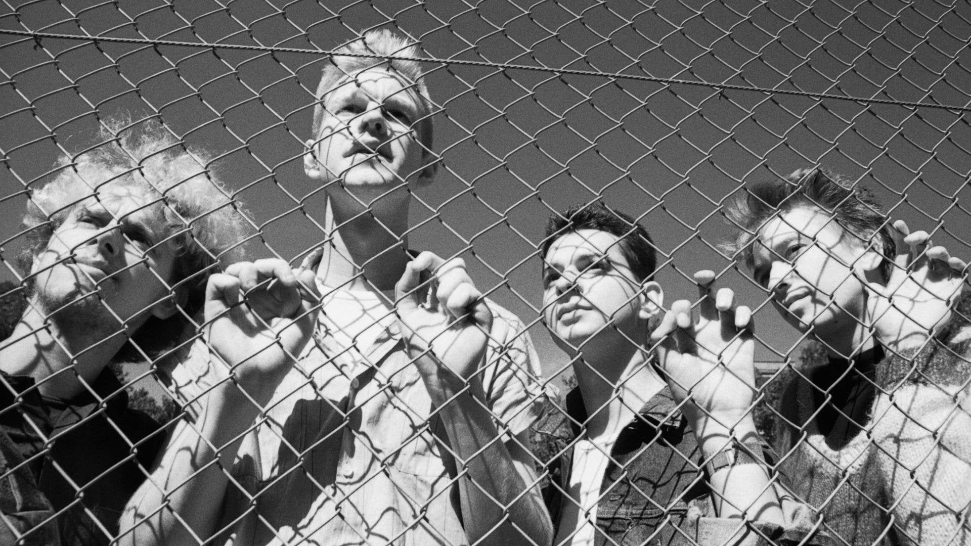 Five individuals, including Alan Wilder, stand behind a chain-link fence, hands raised.