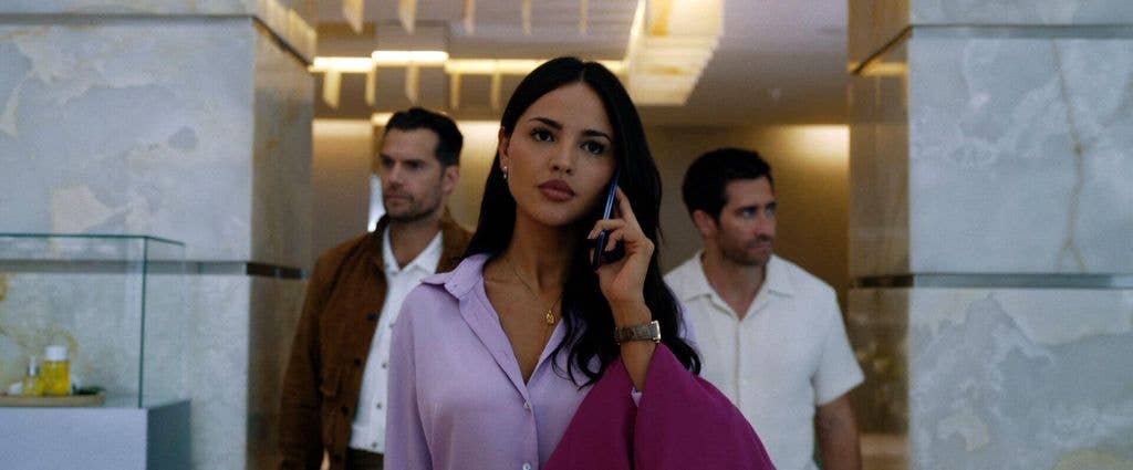 Eiza González Rivera on phone, flanked by two men, one in a brown jacket.