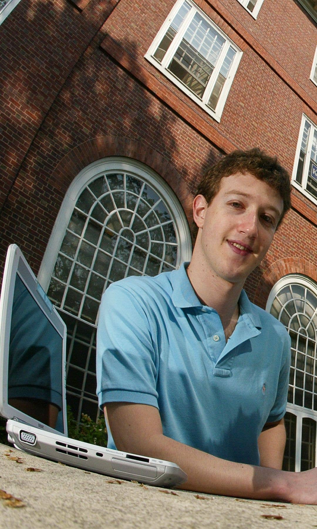 Mark Zuckerberg smiling with a laptop in front of a brick building.