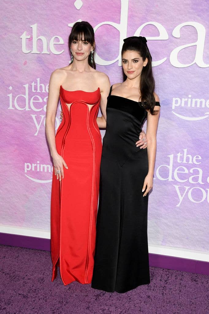 Anne Hathaway wearing a long red dress with her arm around Ella Rubin, wearing a long black dress, in front of The Idea of You sign