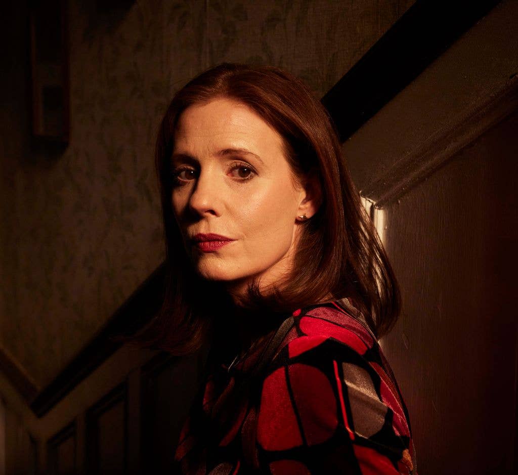 Amy Nuttall stands in a dimly lit hallway, wearing a red and black patterned top.