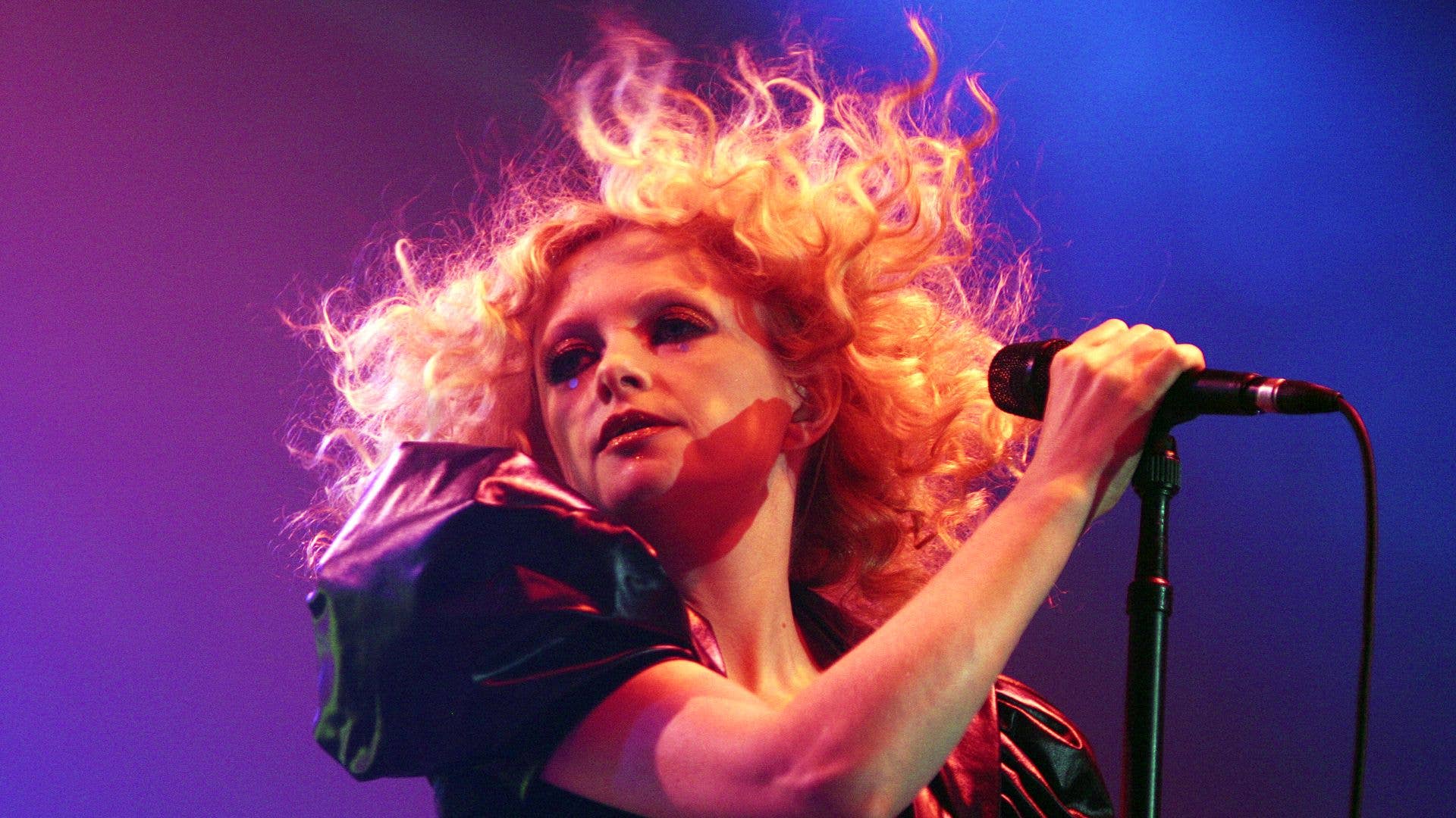 Alison Goldfrapp performing on stage with curly hair, holding a microphone.