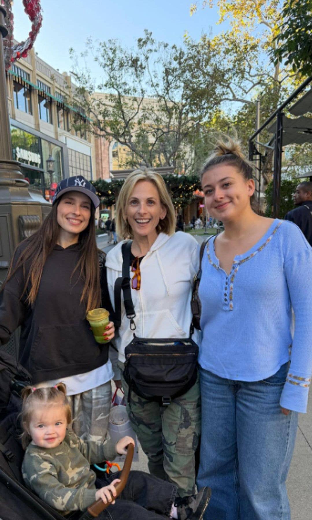 Three women and a child pose for a photo on a city sidewalk. The woman on the left wears a black hoodie and cap, the middle woman in a white jacket is Marlee Matlin, and the woman on the right wears a blue shirt and jeans.