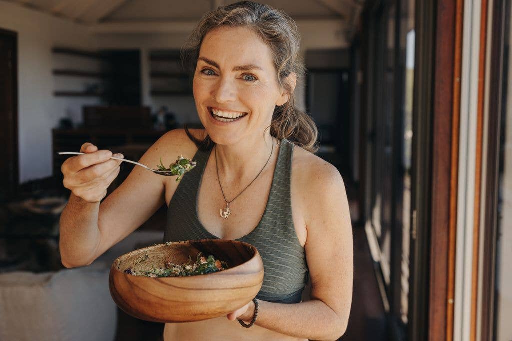 A woman in a gray tank top holds a wooden bowl of food and smiles.