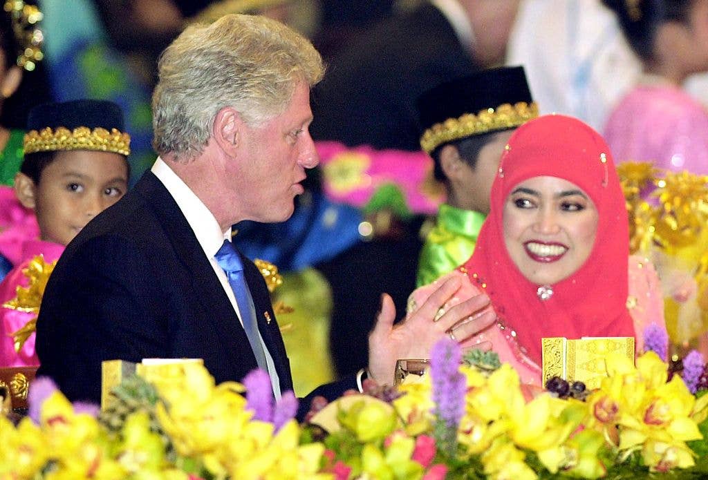 Bill Clinton in a suit chats with Queen Mariam Abdul Aziz in a red hijab at a formal dinner.