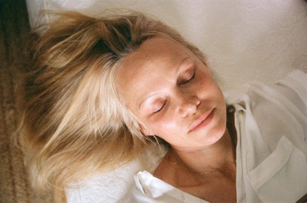 Pamela Anderson lying down with eyes closed, wearing a white shirt.