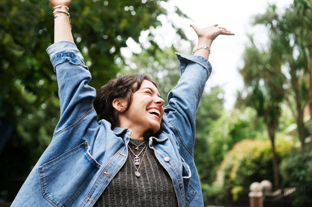 A woman in a denim jacket and gray sweater raises her arms in joy in a park.