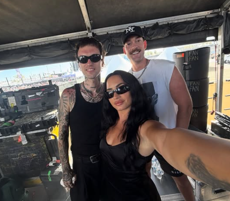 Demi Lovato, with tattoos and sunglasses, takes a selfie with two others in a backstage area.