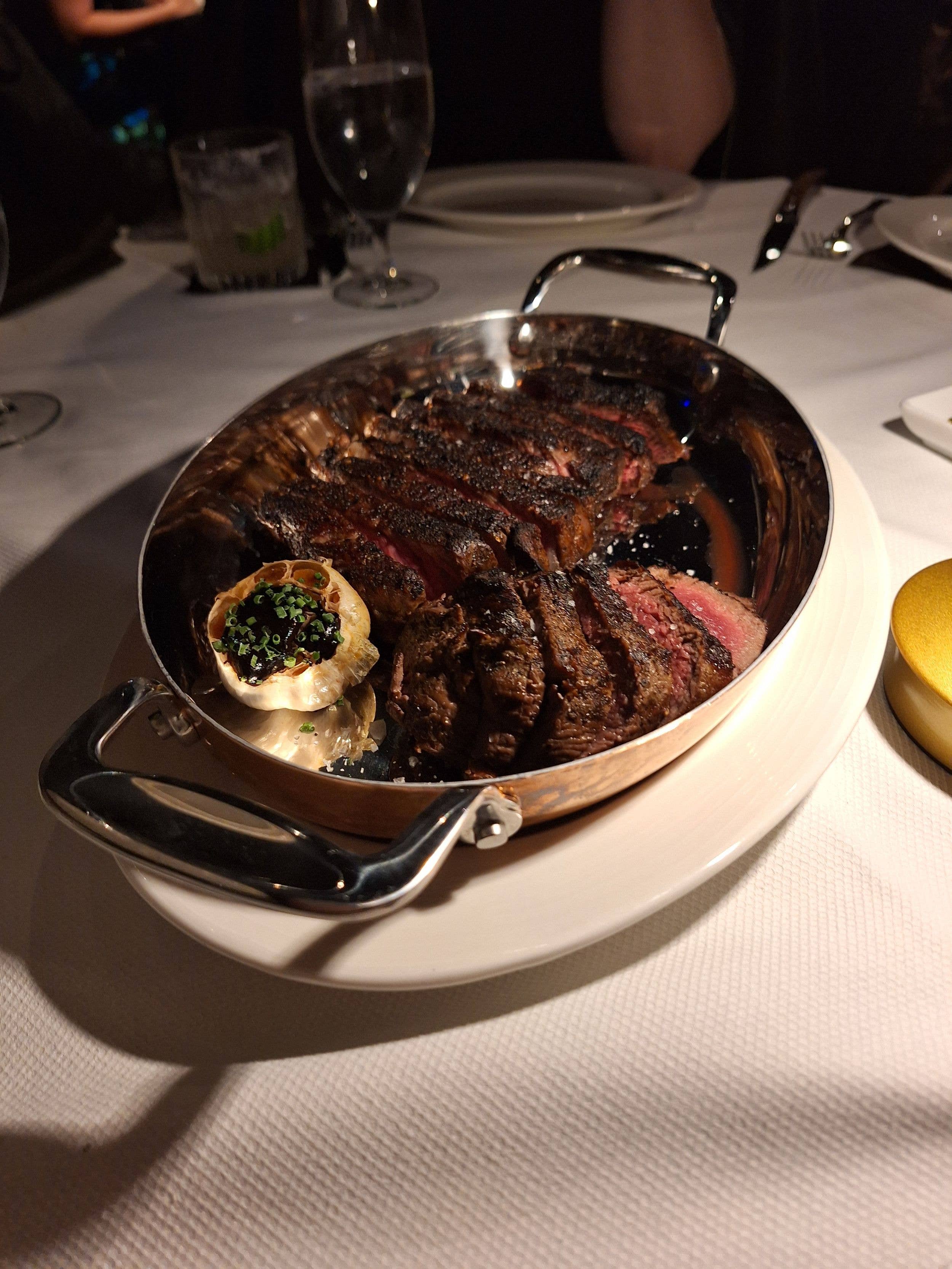 A large, sliced steak in a metal pot on a white plate, with a side dish, on a table with white tablecloth.