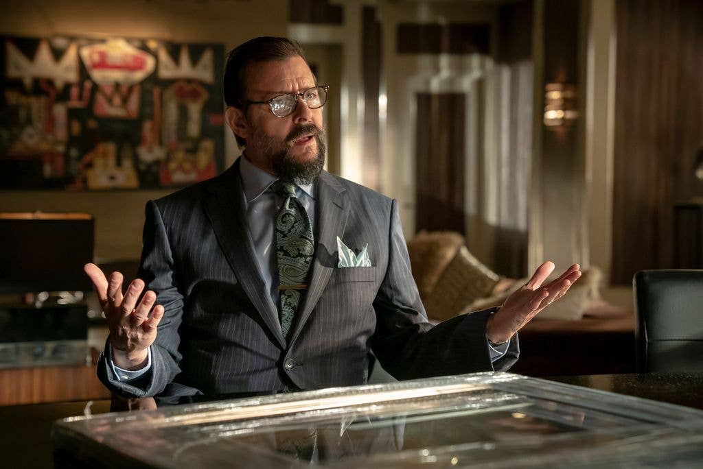 Judd Nelson in a gray suit and green tie, standing with hands open in an office setting.