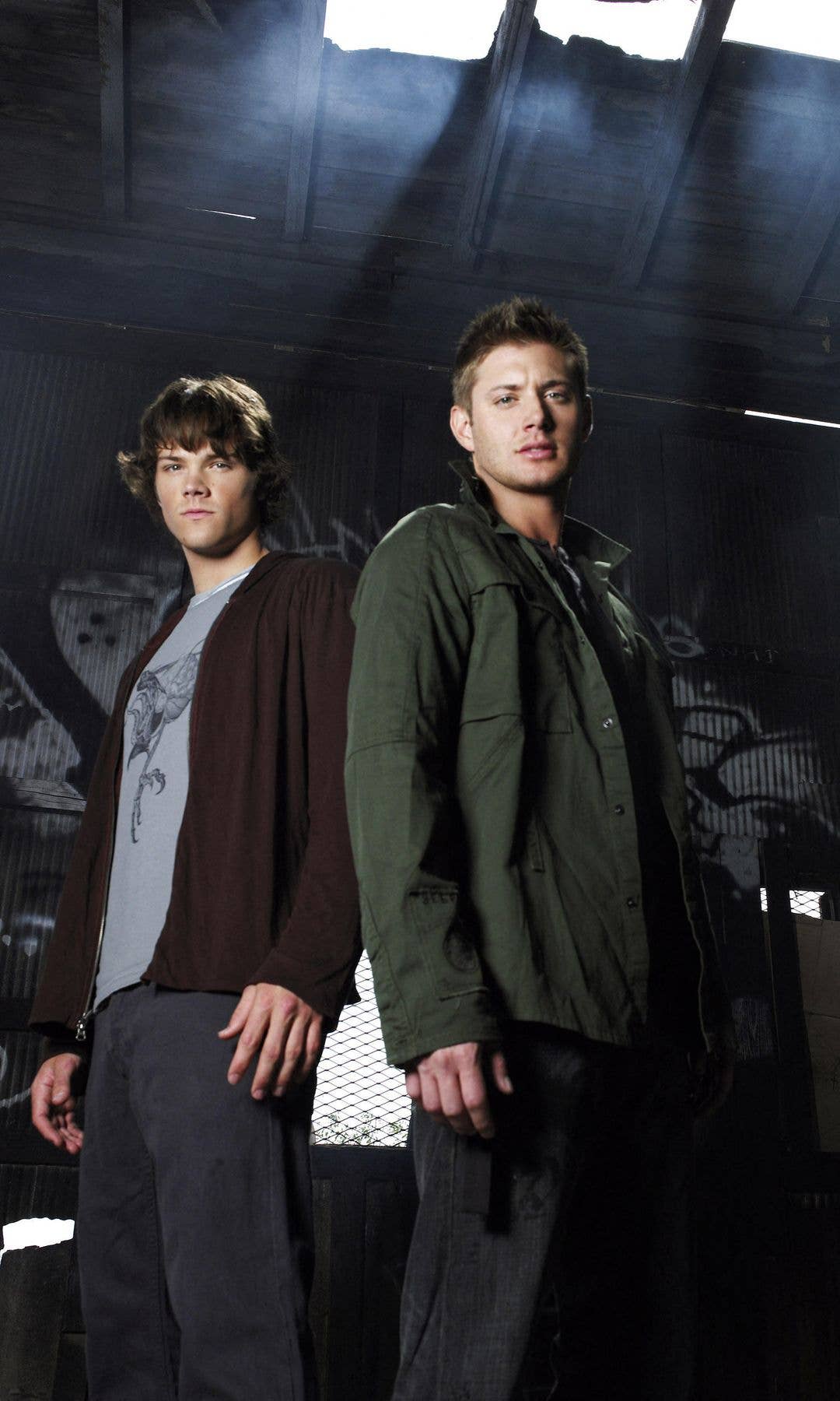 Jensen Ackles and Jared Padalecki stand together in a dark, graffiti-covered room.