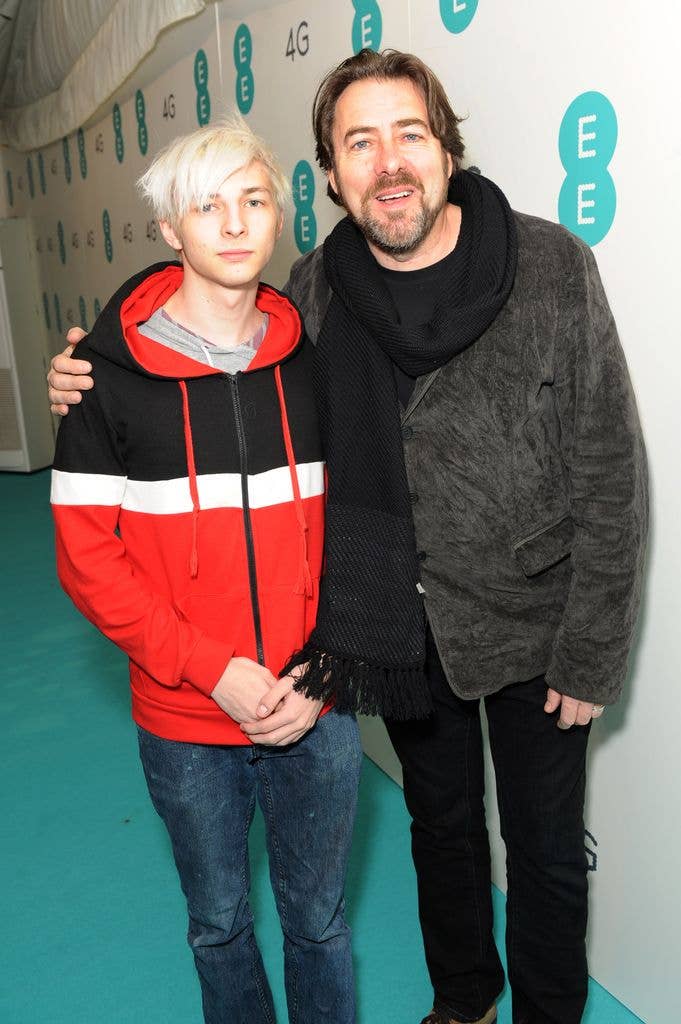 Jonathan Ross, wearing a black scarf and gray jacket, stands with a young man in a red and black hoodie at an event.
