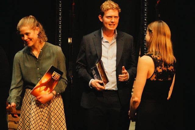 Three people on stage, man in suit holding a book, woman in green blouse holding a book, woman in black dress looking at man.