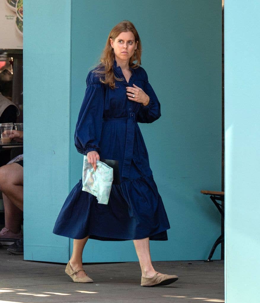 Princess Beatrice in a blue dress, holding a pouch, walking outdoors.