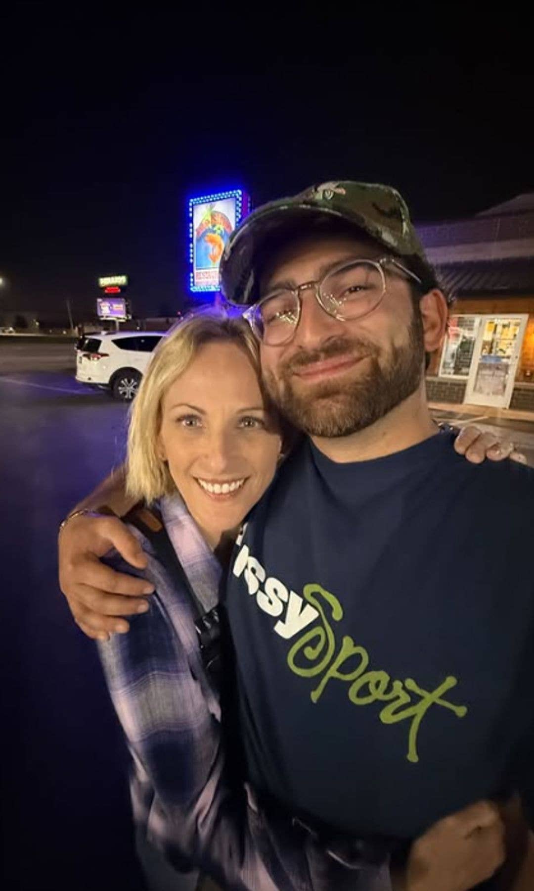 A man and a woman stand close, smiling for a photo at night. The man wears a camo cap and glasses, and the woman has blonde hair and wears a plaid shirt. They are in front of a brightly lit sign.