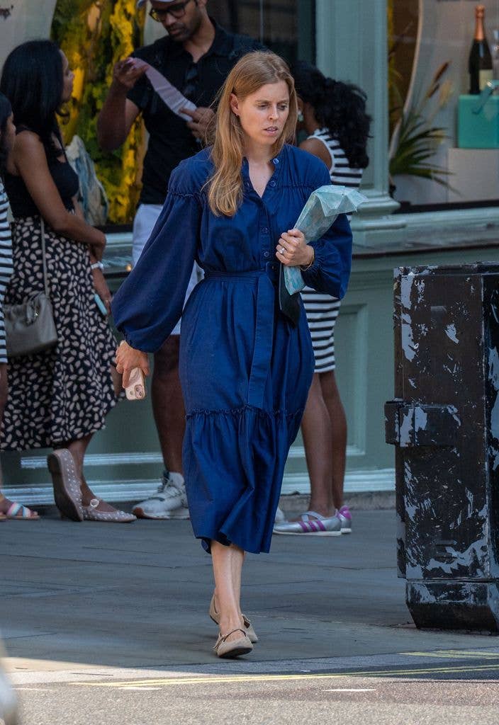 Princess Beatrice in a blue dress, holding a bag, walking on a sidewalk.