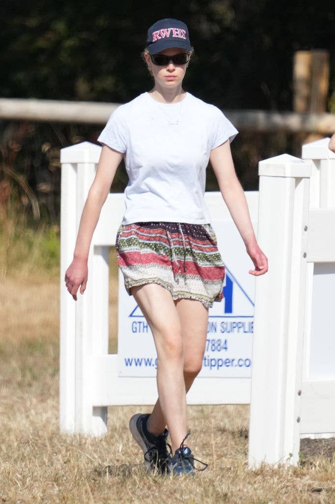 A woman in a white t-shirt, patterned shorts, and a cap walks on grass.