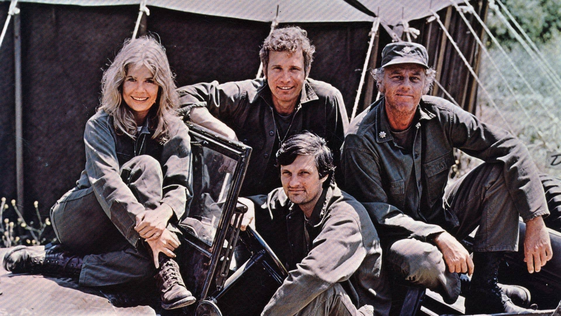 Four individuals in military attire pose on a jeep. Loretta Swit sits on the hood, smiling.