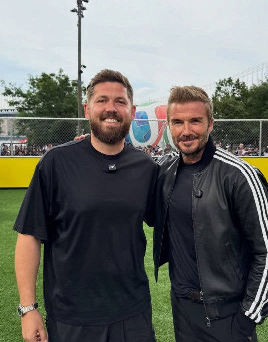 David Beckham stands next to John Nellis, both smiling, on a green field.