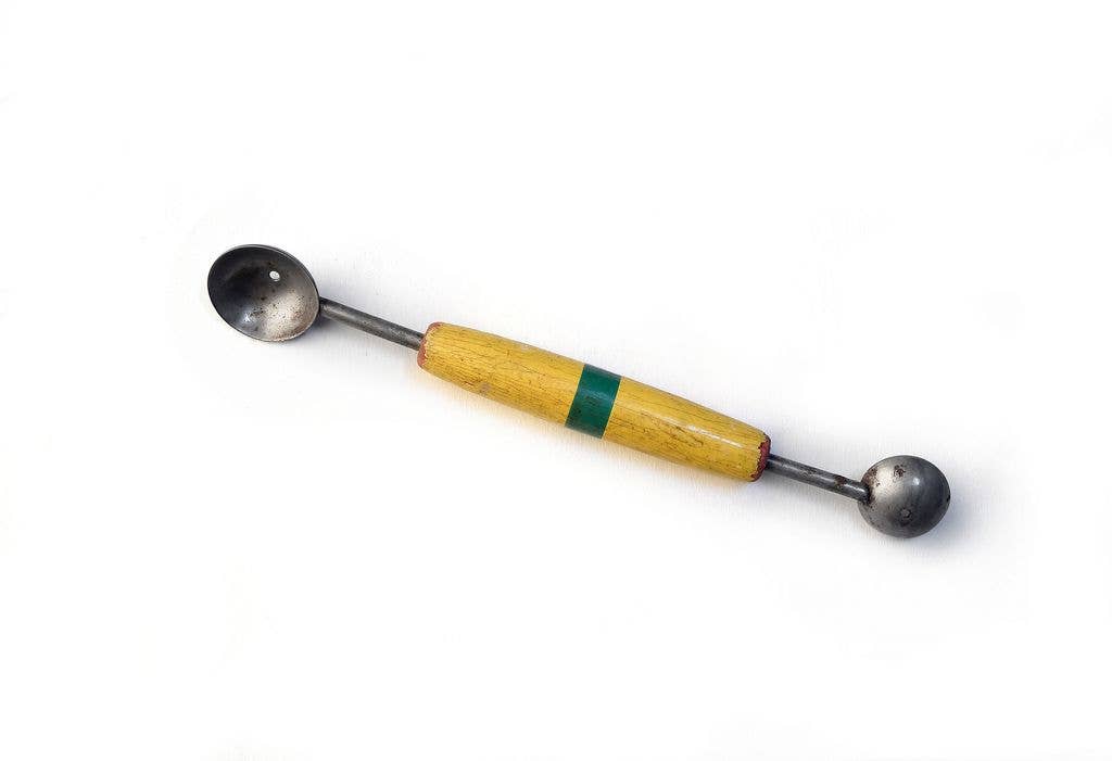 A vintage metal melon baller with a yellow wood handle featuring a green stripe, one end shaped as an oval scoop and the other as a round scoop, against a white background.